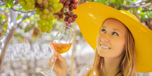 Una joven disfrutando de una experiencia de enoturismo en una bodega española. Se encuentra entre viñedos verdes, sosteniendo una copa de vino mientras sonríe, con la bodega tradicional y un cielo azul claro de fondo, simbolizando la conexión entre el turismo, la cultura y la pasión por el vino en España.